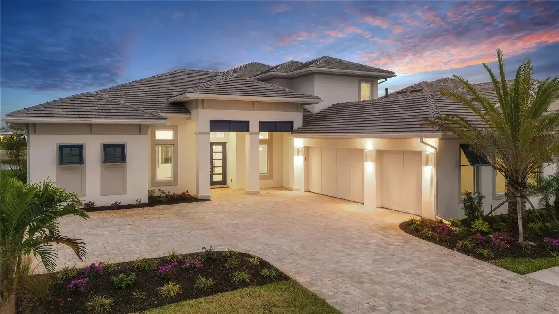 Elegant contemporary home with a spacious driveway, lush landscaping, and dramatic sunset sky, highlighting modern architecture and inviting ambiance.